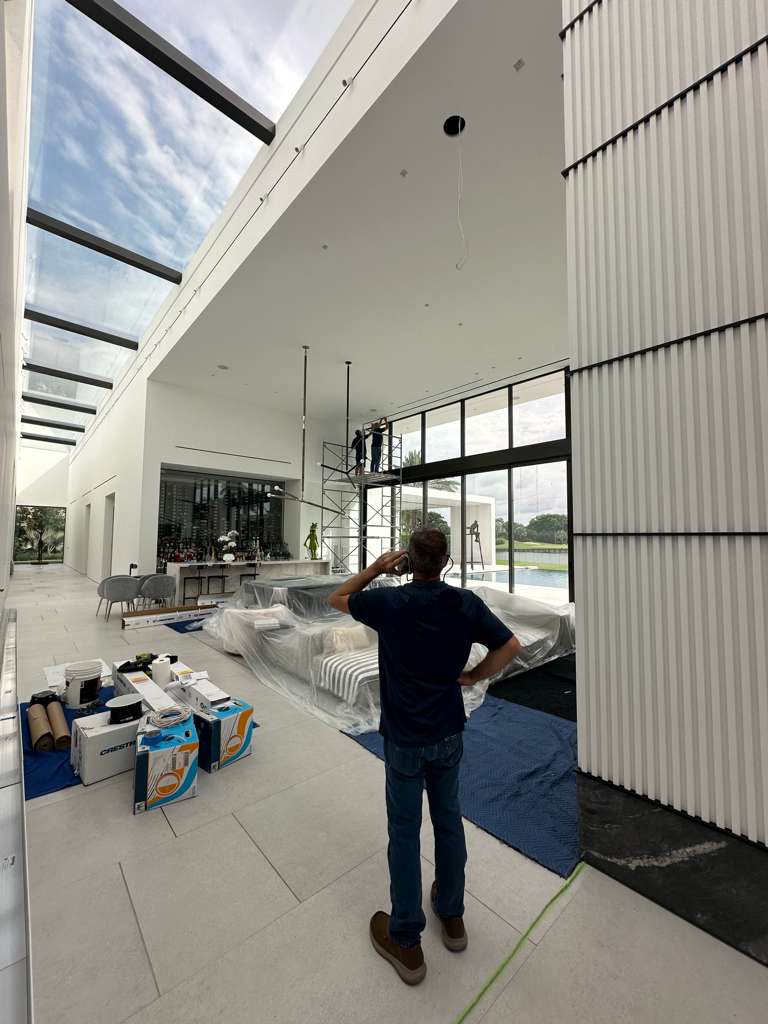 Man observing a technician on scaffolding installing technology in a modern, spacious living area with large windows, surrounded by installation materials and covered furniture, highlighting the integration of smart technology in elegant home design.